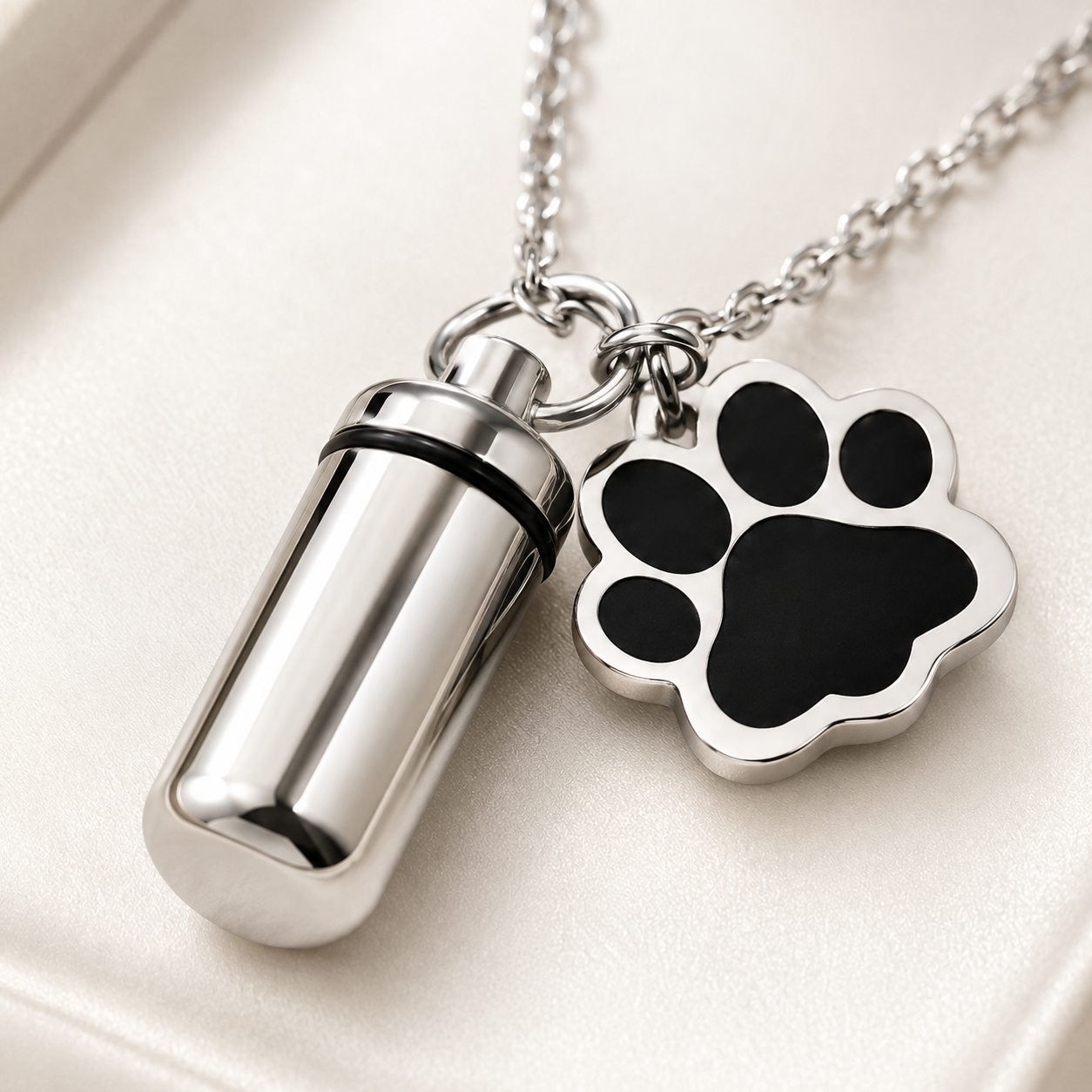 Close-up of polished silver pet memorial keepsake pendant and black paw-print charm on an ivory surface.